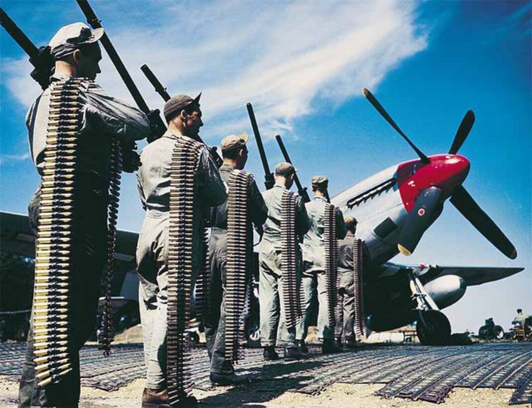 Six crew members lined-up holding ammo next to a P-51 Mustang