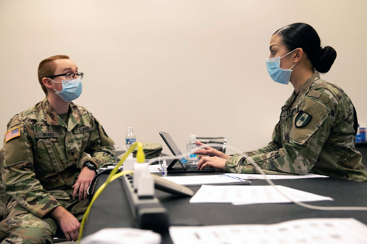 A woman wearing a military uniform sits behind a desk and types on a laptop while talking with a man in a military uniform who is seated opposite her. Both are wearing face masks.