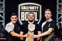 two white males in black, white and yellow tshirts hold a brown esports trophy with male with beard in black shirt stands in the center. A sign tthat says Call of Duty Endowment Bowl is in the background.