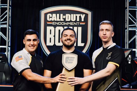 two white males in black, white and yellow tshirts hold a brown esports trophy with male with beard in black shirt stands in the center. A sign tthat says Call of Duty Endowment Bowl is in the background.