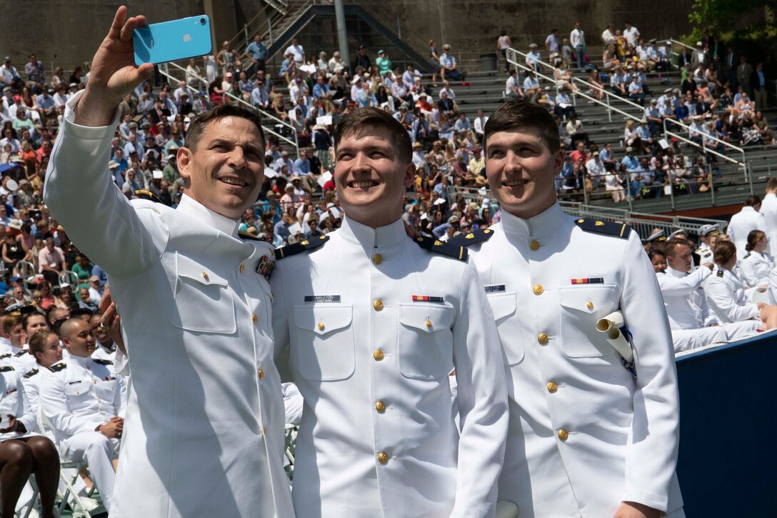 A father and his twin sons, wearing dress white uniforms, smile for a selfie.