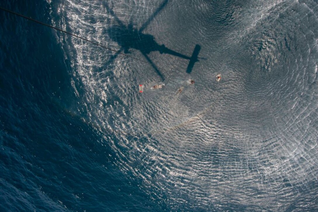 A group of sailors swim in open water with the shadow of a helicopter above them visible in the sea.