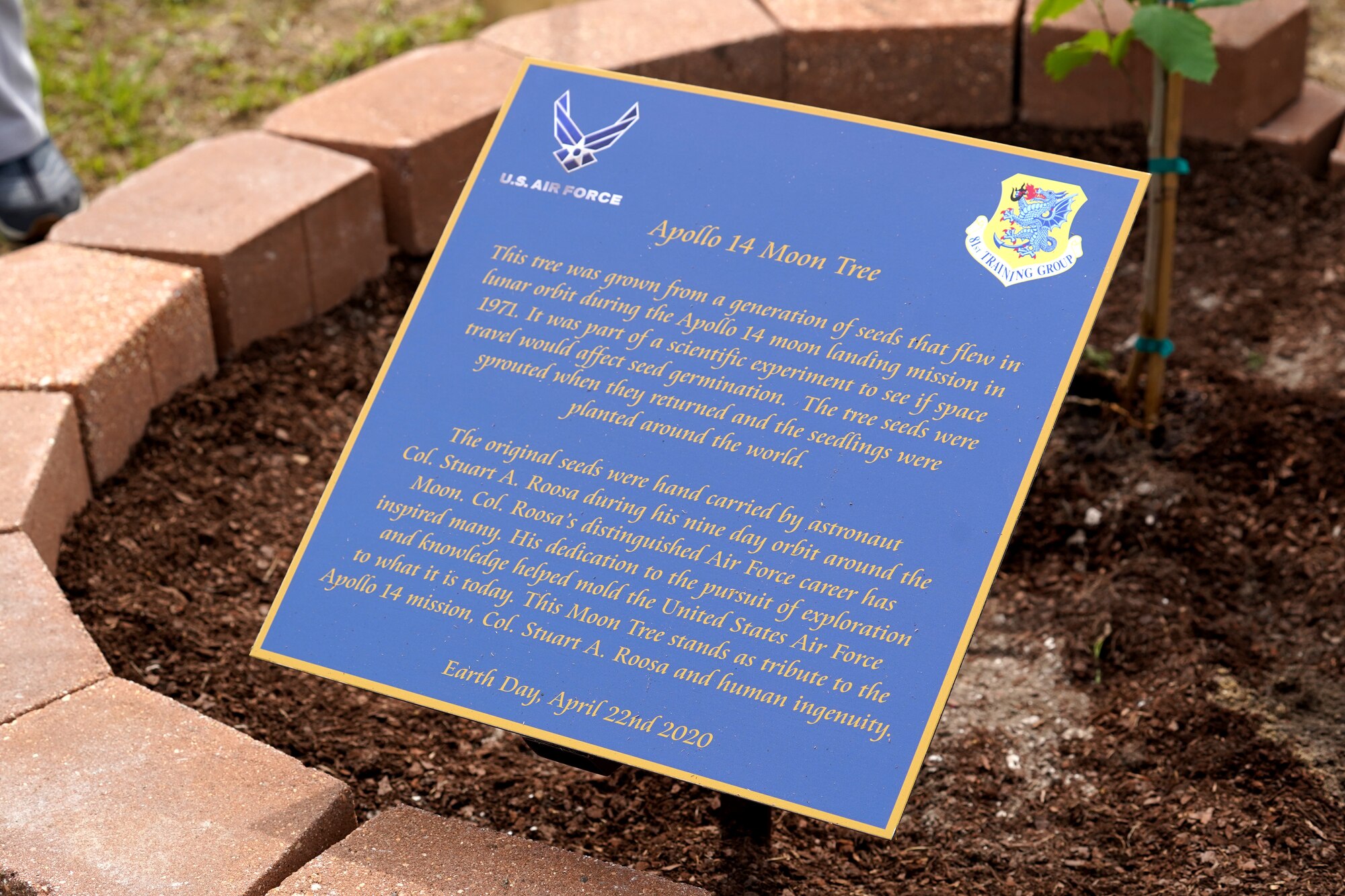 A sign is displayed during a tree-planting ceremony at Keesler Air Force Base, Mississippi, April 22, 2020. The tree was grown from a generation of seeds that flew with astronaut Col. Stuart A. Roosa as an experiment on mission Apollo 14. The tree was planted on Earth Day as a tribute to Apollo 14, Roosa, and human ingenuity. (U.S. Air Force photo by Airman 1st Class Seth Haddix)
