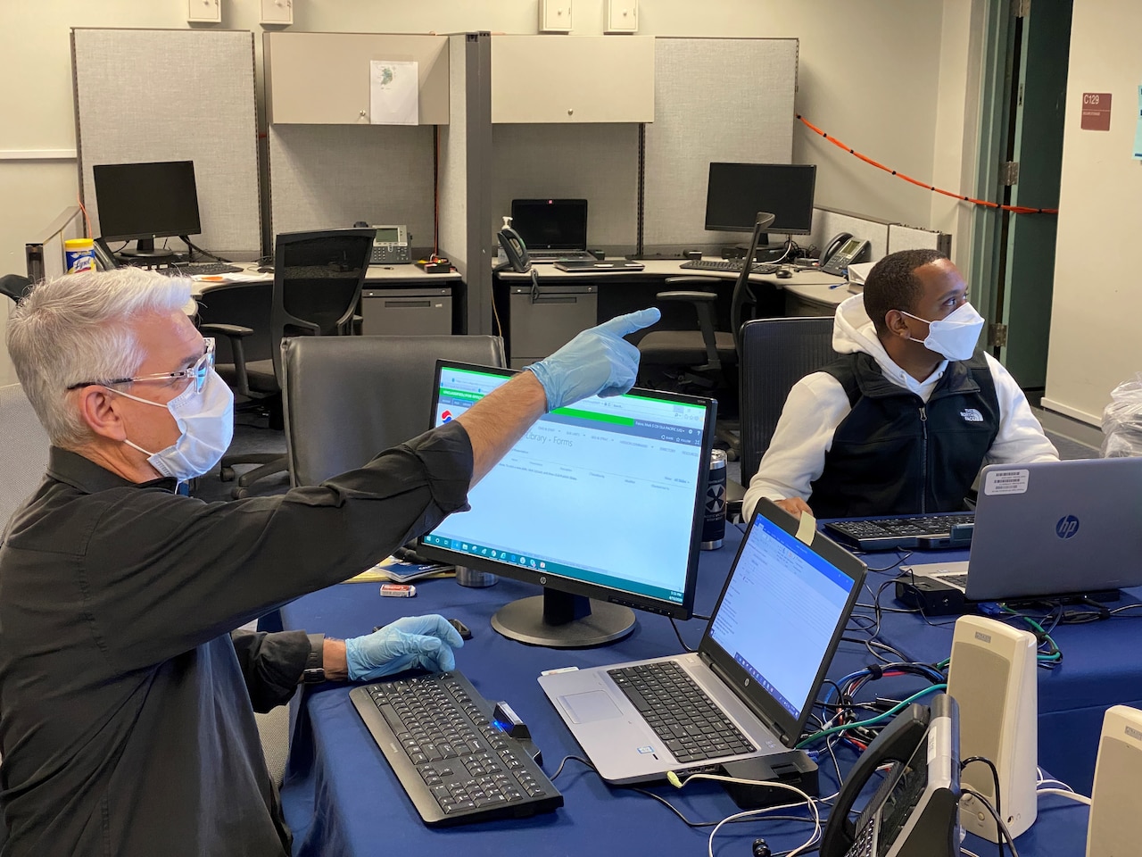 Two men sit at a table with computers next to each other while they both look at what one man is pointing at out of frame.