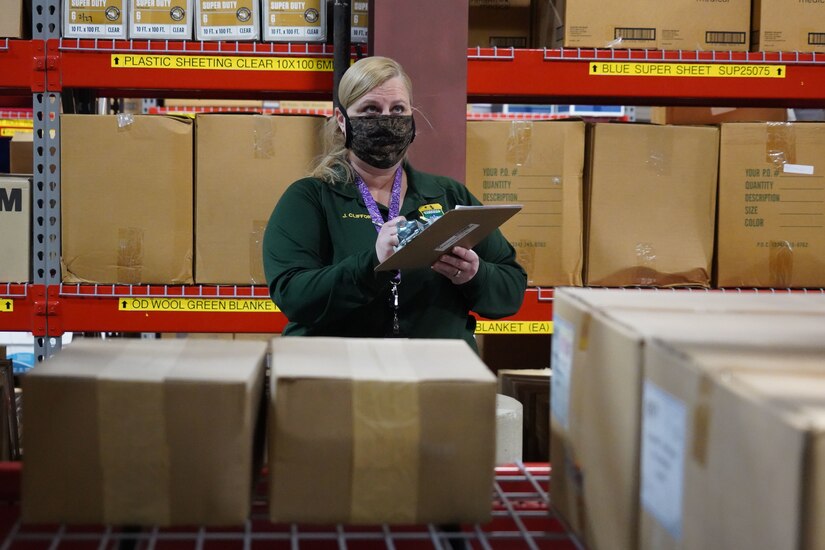 A woman wearing a mask and holding a clipboard stands amid a lot of boxes.