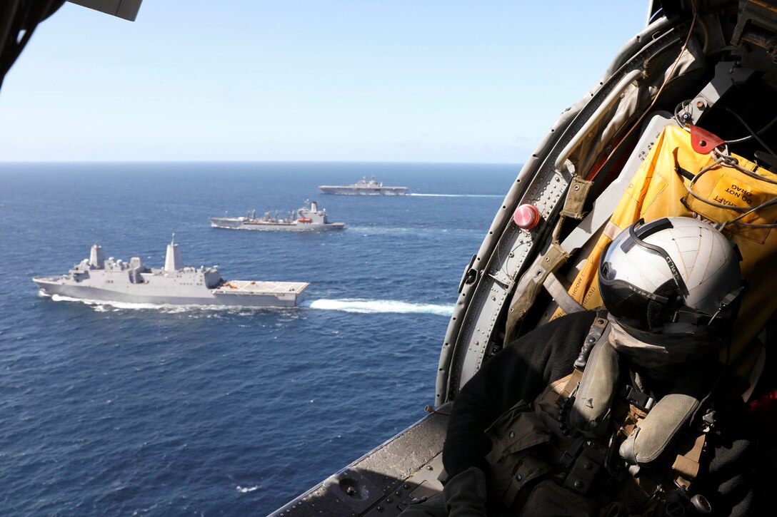 A sailor flies in a helicopter while three ships sail below.