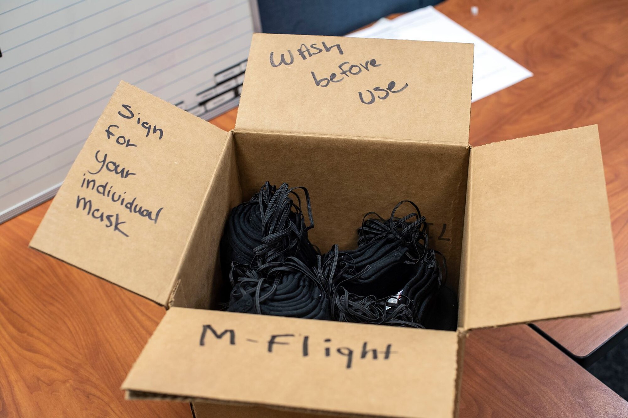 A cardboard box with the phrases "wash before use," "Sign for your individual mask," and "M-Flight" written on it sits on a wooden table with black face masks in it