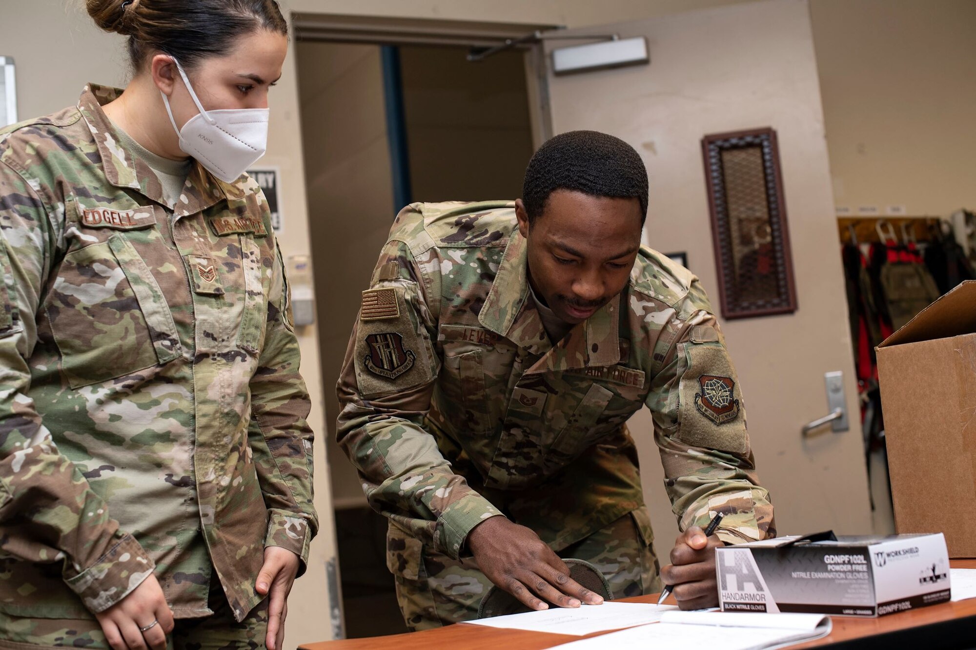 A female Airmen with a white face mask on supervises a male Airmen signing a document in front of a box full of black face masks