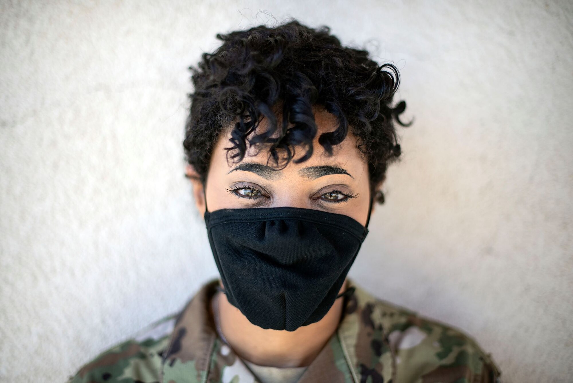 A female Airman in uniform looks directly at the camera with a black face mask on. It's sunny.