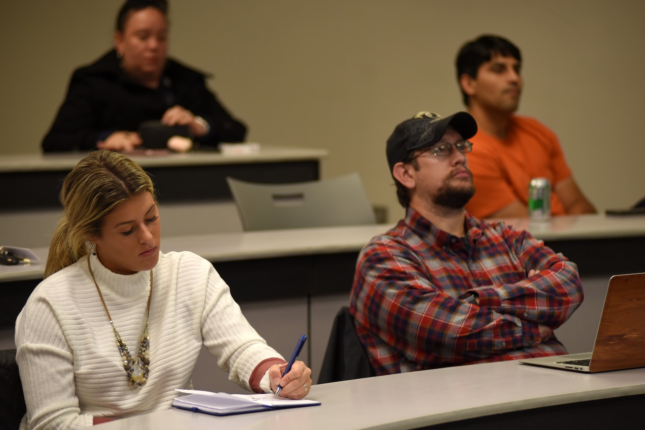 Men and women seated at tables in rows listen and take notes in a college classroom setting.