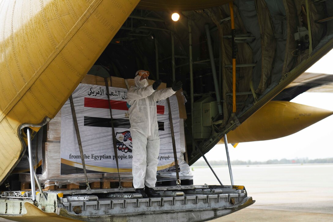 A service member stands at the back of an aircraft with boxes stacked behind.