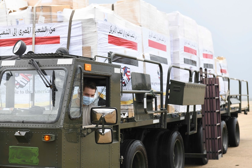 A service member drives a large truck with boxes in the back.