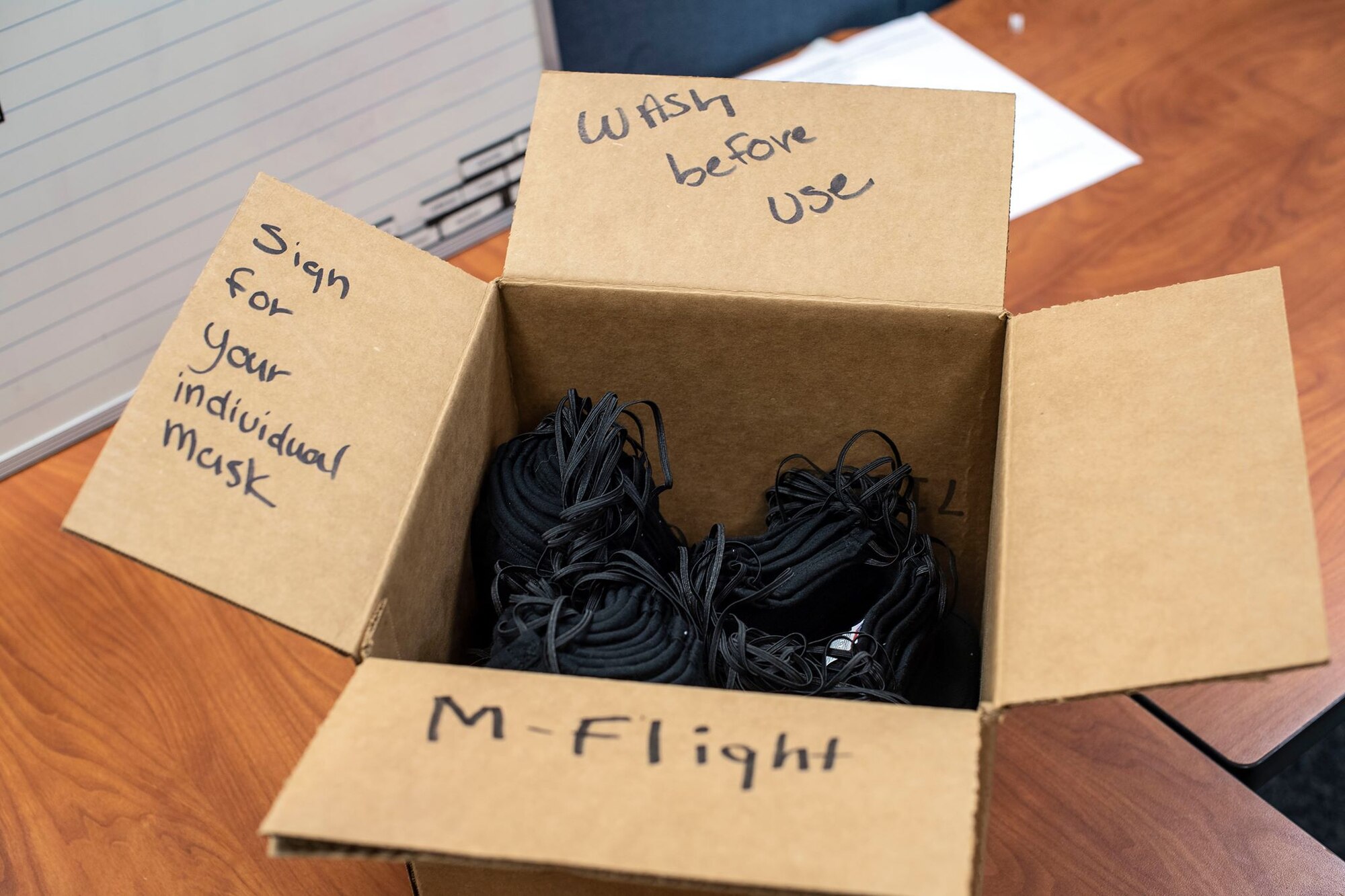 A cardboard box with the phrases "wash before use," "Sign for your individual mask," and "M-Flight" written on it sits on a wooden table with black face masks in it