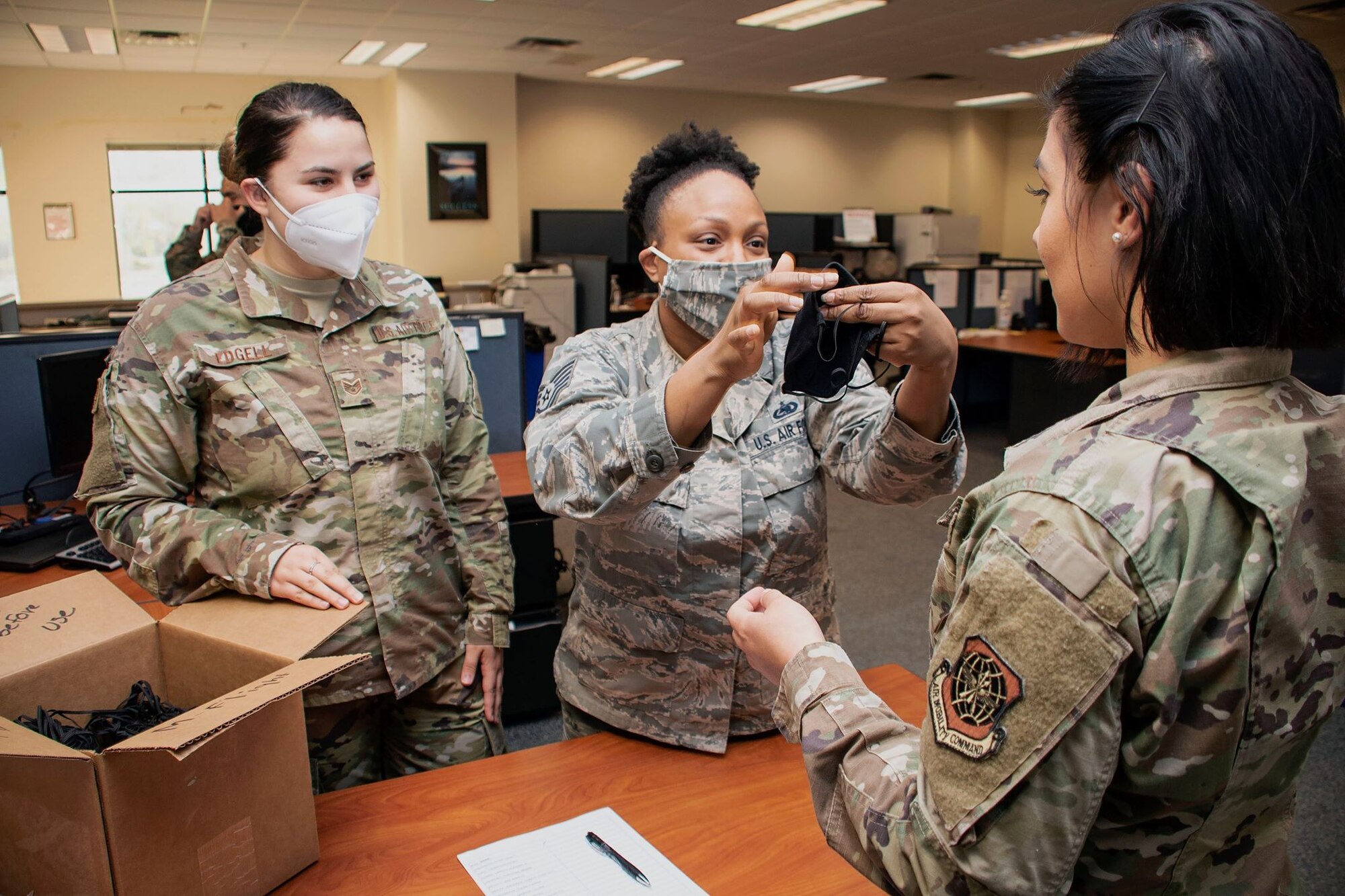 Two female Airmen, one with a white face mask and one with a camo face mask on, instructs a third face mask-less Airmen on how to don one.