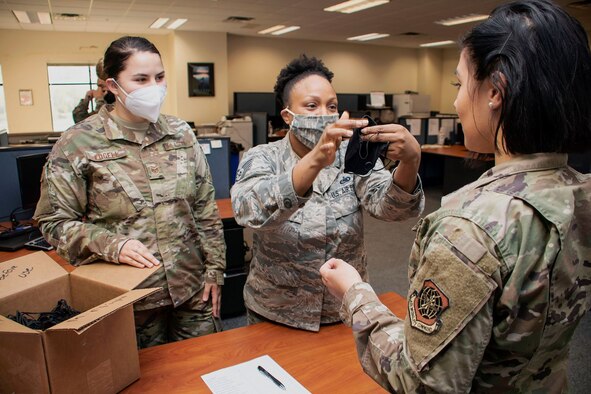 Two female Airmen, one with a white face mask and one with a camo face mask on, instructs a third face mask-less Airmen on how to don one.