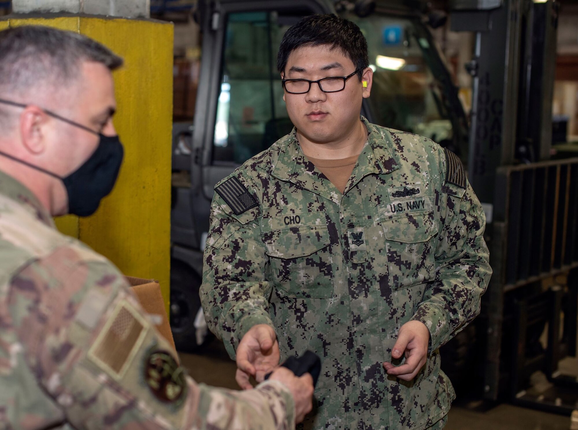 A male Airman with a black face mask on hands off another black face mask to a male Sailor in a warehouse-type environment