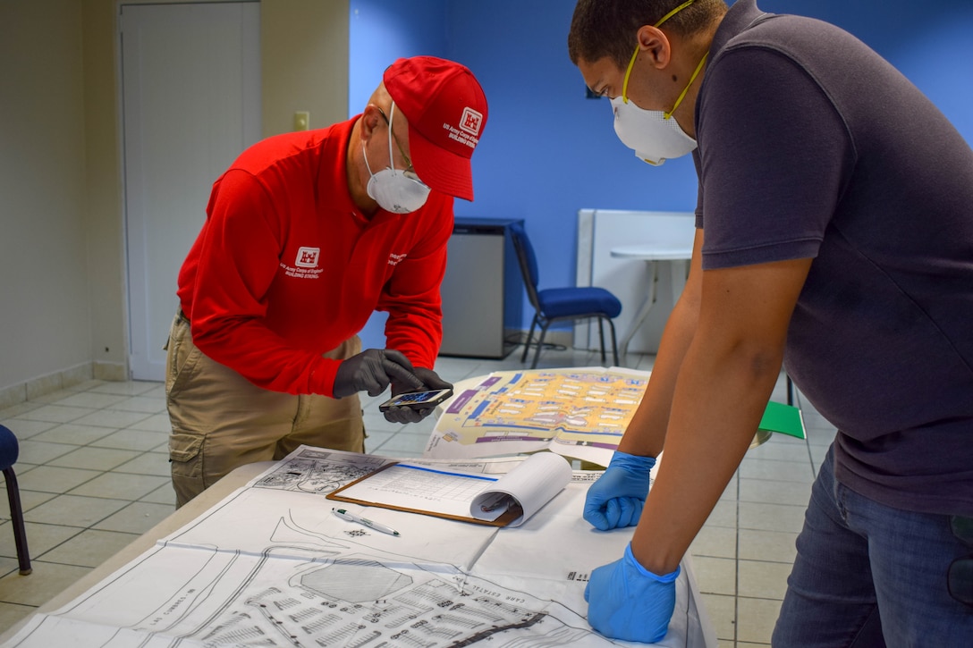 Ramon Collazos wearing a red USACE shirt imputing numbers on a calculator while looking at map layout on the table as Alejandro Feliz  observes.
