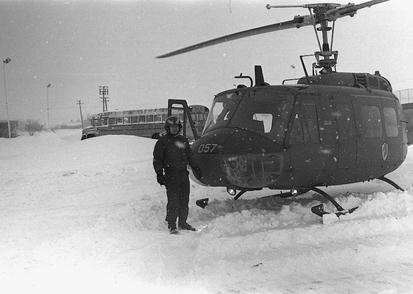 A man in military pilot clothing stands outside a helicopter in the snow. There's a school bus in the background.