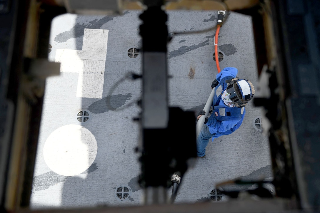 A sailor underneath a helicopter holding a large hook.