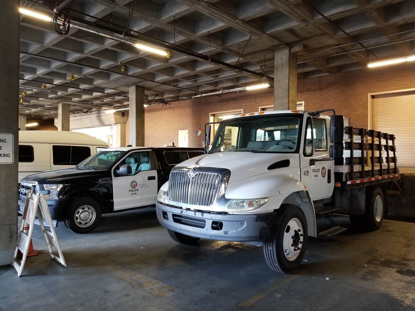 White flatbed truck with brown stakes parked in a parking structure next to a pickup and an a frame sign