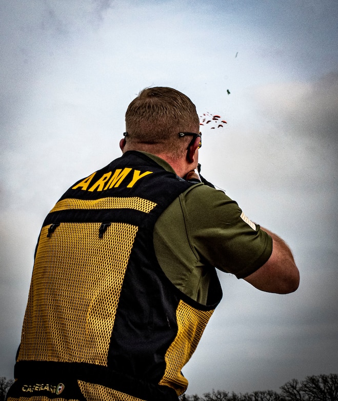 white male in green shirt and yellow and black vest wearing shoots a clay pigeon