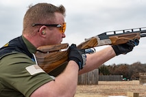 white male in green shirt and black vest wearing orange lens glasses, shooting gloves and hearing protection holds a rifle.
