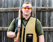 white male in green shirt and yellow and black vest wearing orange lens glasses, shooting gloves and hearing protection holds a rifle.