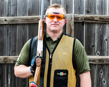 white male in green shirt and yellow and black vest wearing orange lens glasses, shooting gloves and hearing protection holds a rifle.