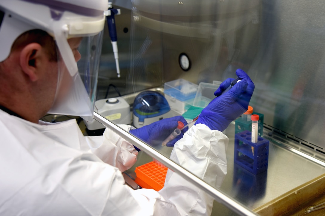 A medical operations officer in a protective suit and gloves sitting in a lab looks at a COVID-19 test vial.