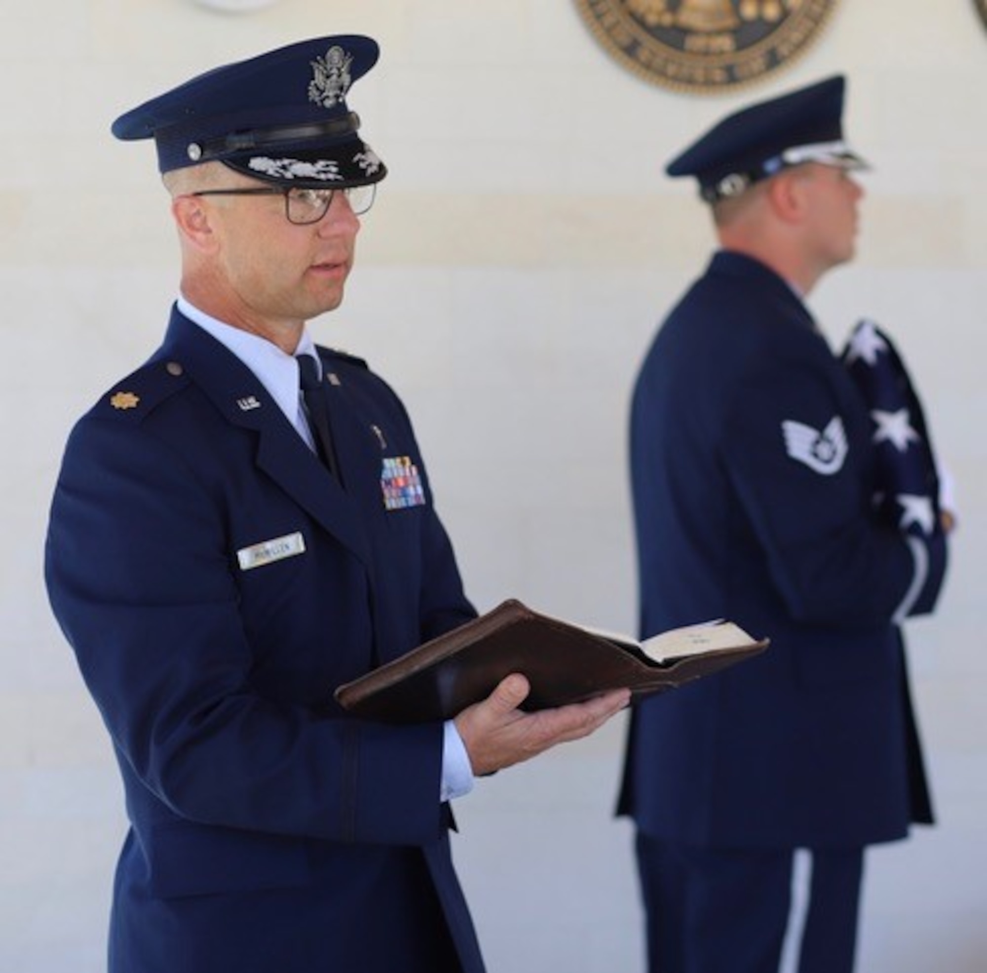 Chaplain in military uniform holds bible with an honor guard member holding an American flag in the background.