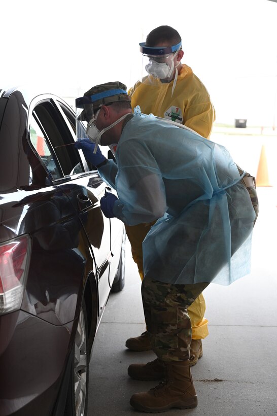 Photo of two North Dakota Air National Guard medical personnel wearing personal protective equipment as they gather COVID-19 swab samples using a q-tip from volunteers driving in a car in the parking lot of the FargoDome, Fargo, N.D., April 25, 2020.