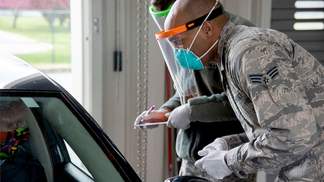 A black soldier wearing a mask and medical mask over his nose and mouth and white gloves talks to a man also wearing a mask sitting in a car.