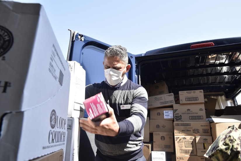 A man wearing a face mask prepares to load boxes into the open back of a van behind him. The van already contains several boxes.