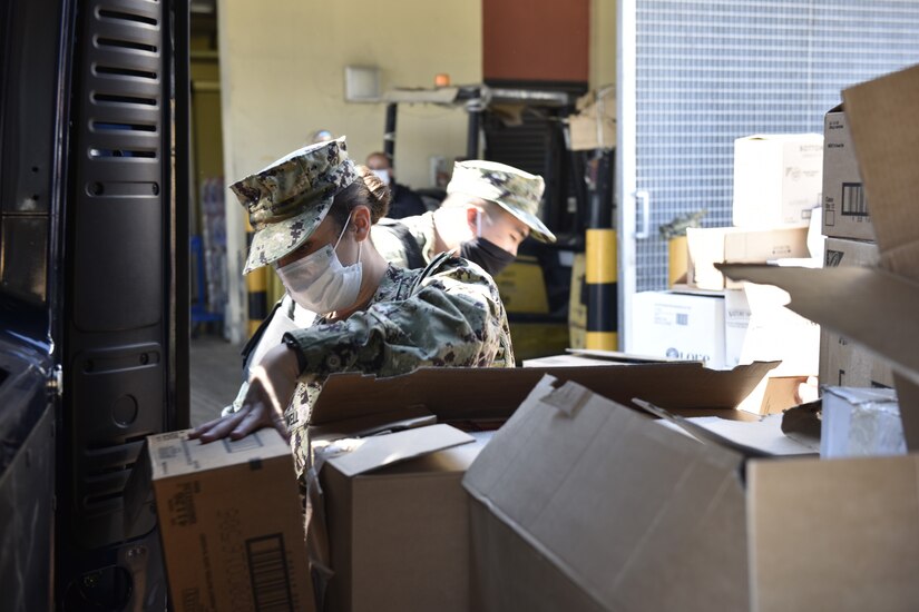 Two sailors in uniform, both wearing face masks, load boxes into a van.