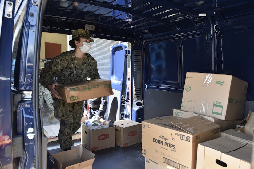 A sailor in uniform and wearing a face mask loads a box into the back of a van.