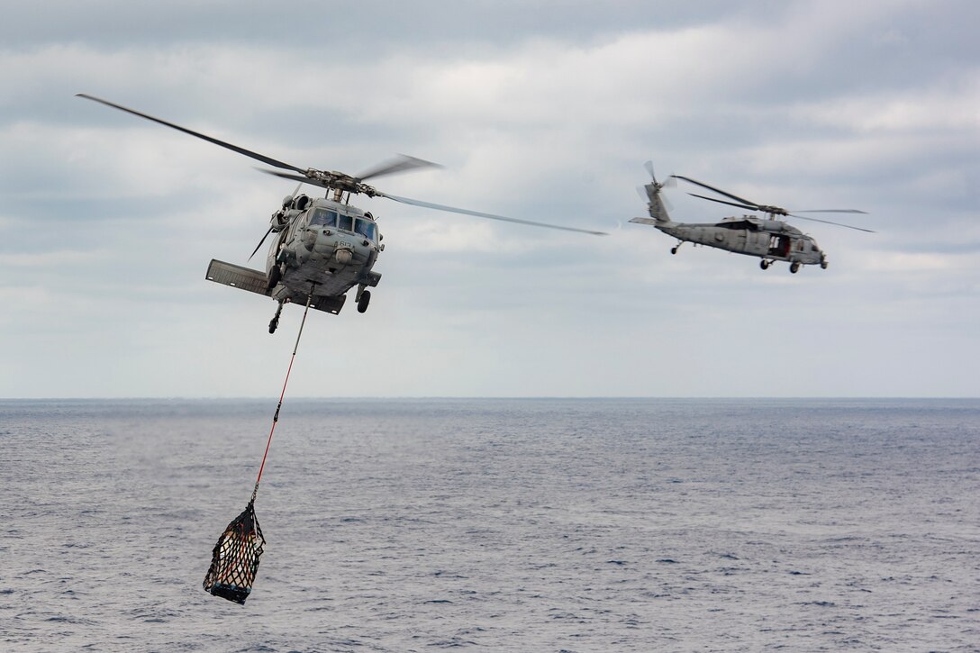 Two Navy helicopters fly near the ocean.