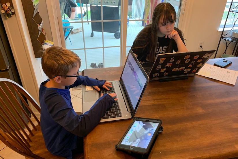Two children sit at a kitchen table and operate laptop computers.