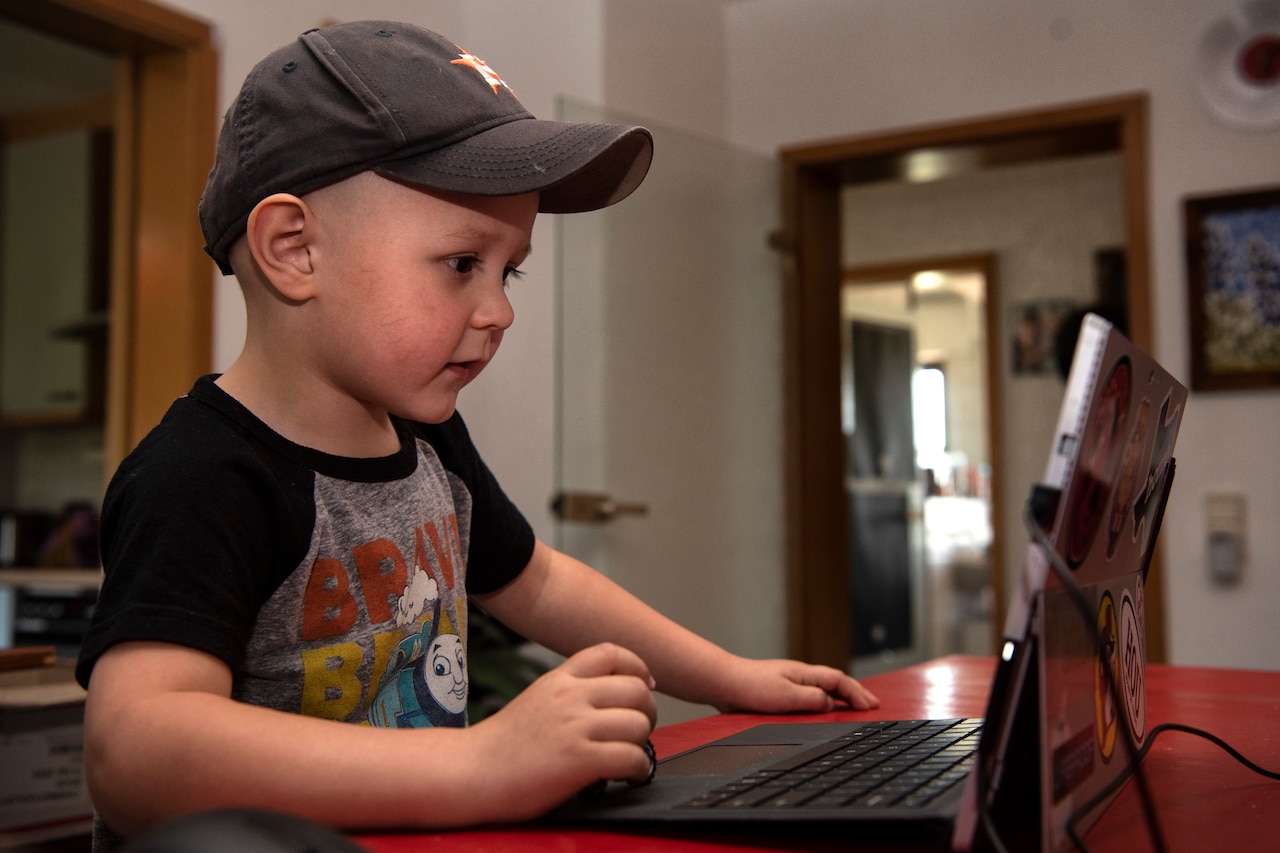 A small boy wearing a baseball cap and Thomas the Tank Engine T-shirt looks at a computer screen.