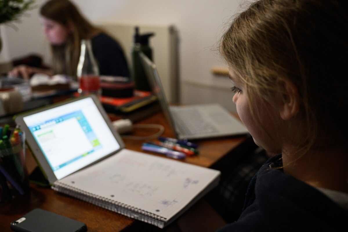 A young girl sits at a kitchen table and looks at a laptop computer screen.  In the background, a young woman also looks at a computer screen.