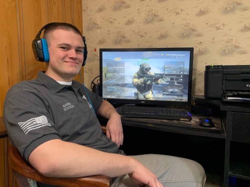 white male in gray and white shirt and blue and black headset sits in front of a computer with a video game Soldier on the screen.