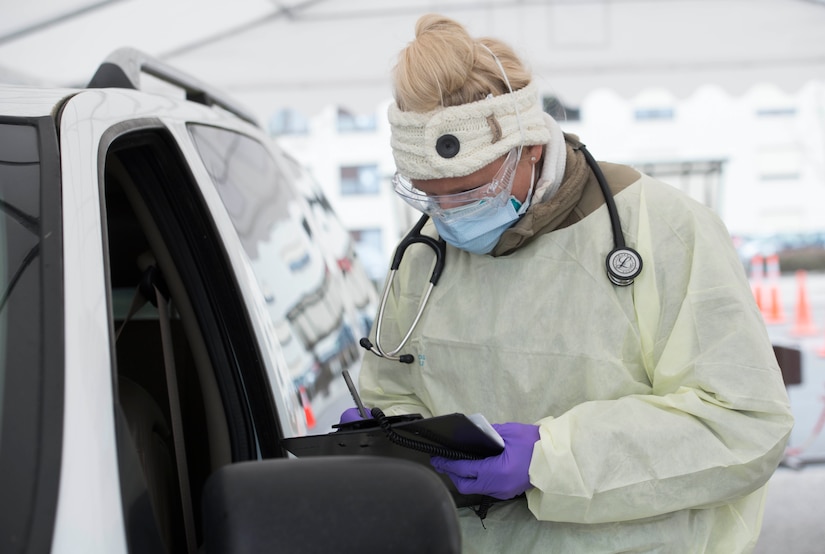 A woman dressed in personal protective equipment takes information from a motorist through a car window.