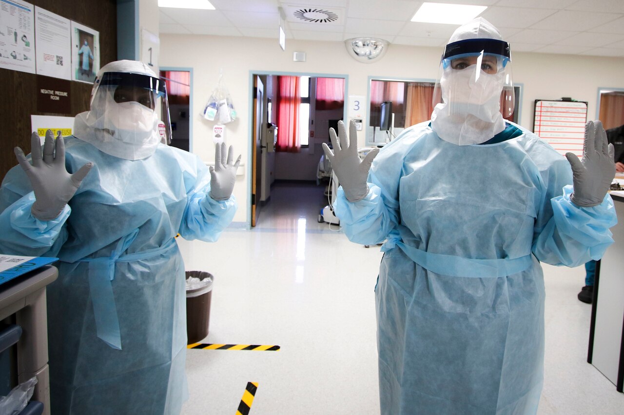 Two people dressed head to toe in personal protective equipment hold up their gloved hands.
