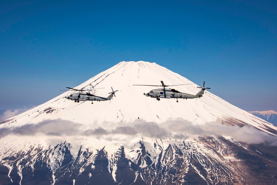 Two helicopters fly in the sky; a mountain can be seen in the background.