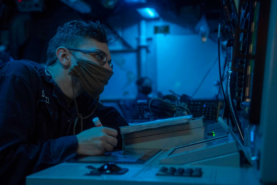 A sailor sits at a desk in front of monitors surrounded by blue light.