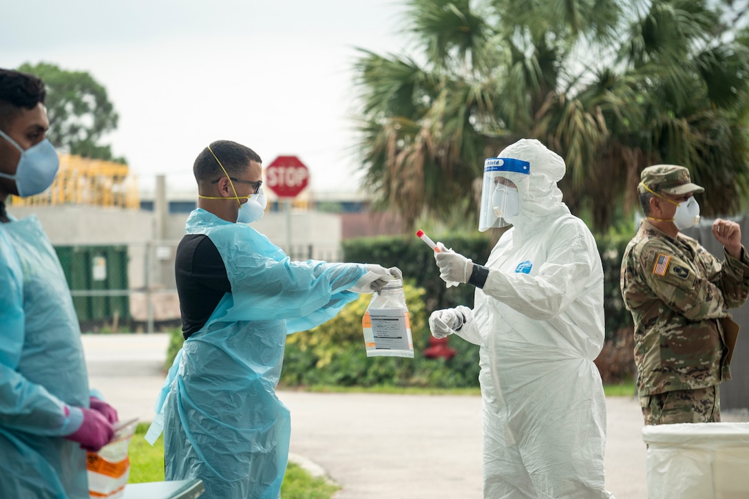 A soldier in protective gear hands a vial to another soldier in protective gear holding out a plastic bag outside.