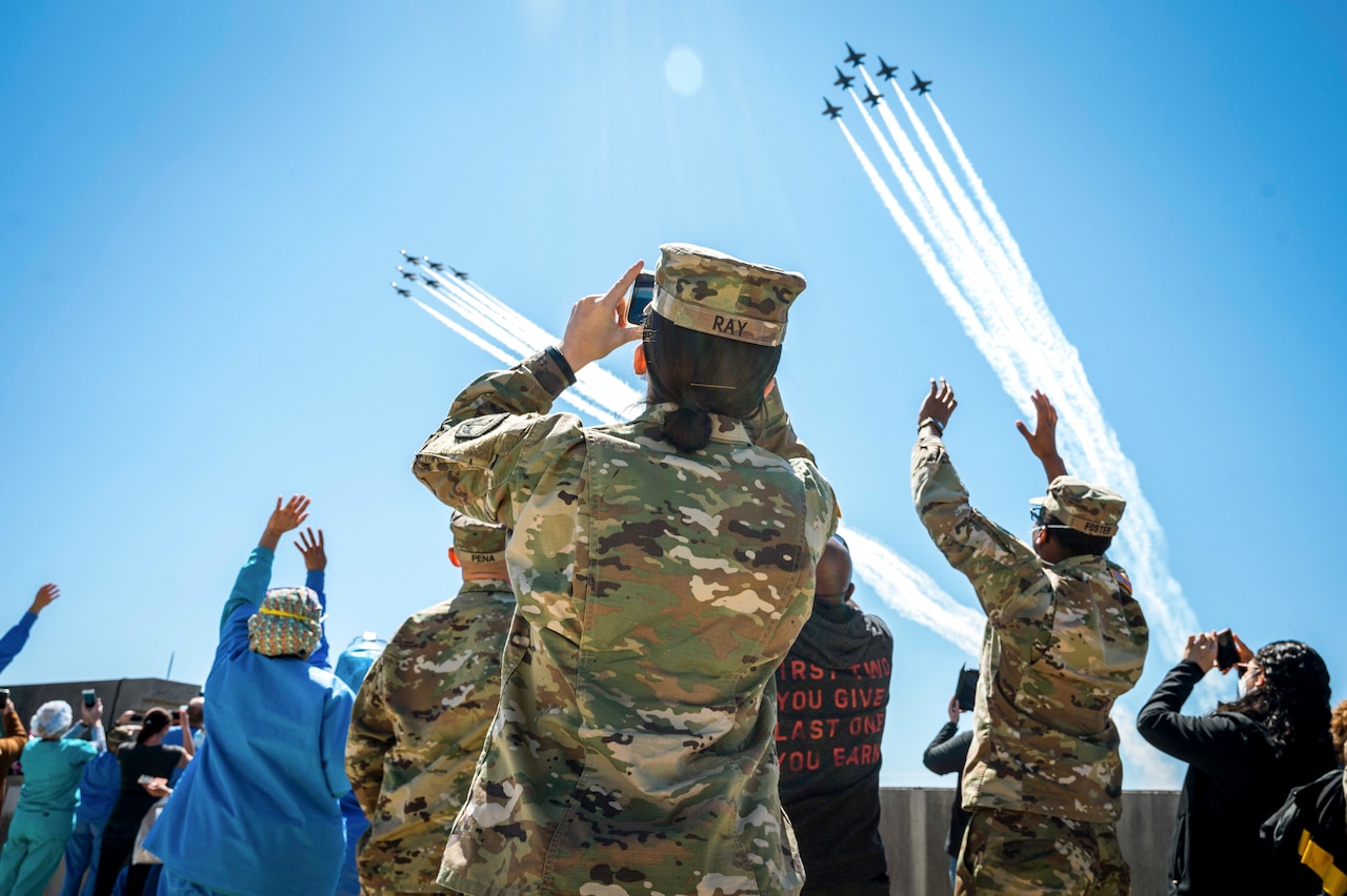 A soldier standing in a crowd of waving people wearing masks takes a photo of two jet formations flying overhead.