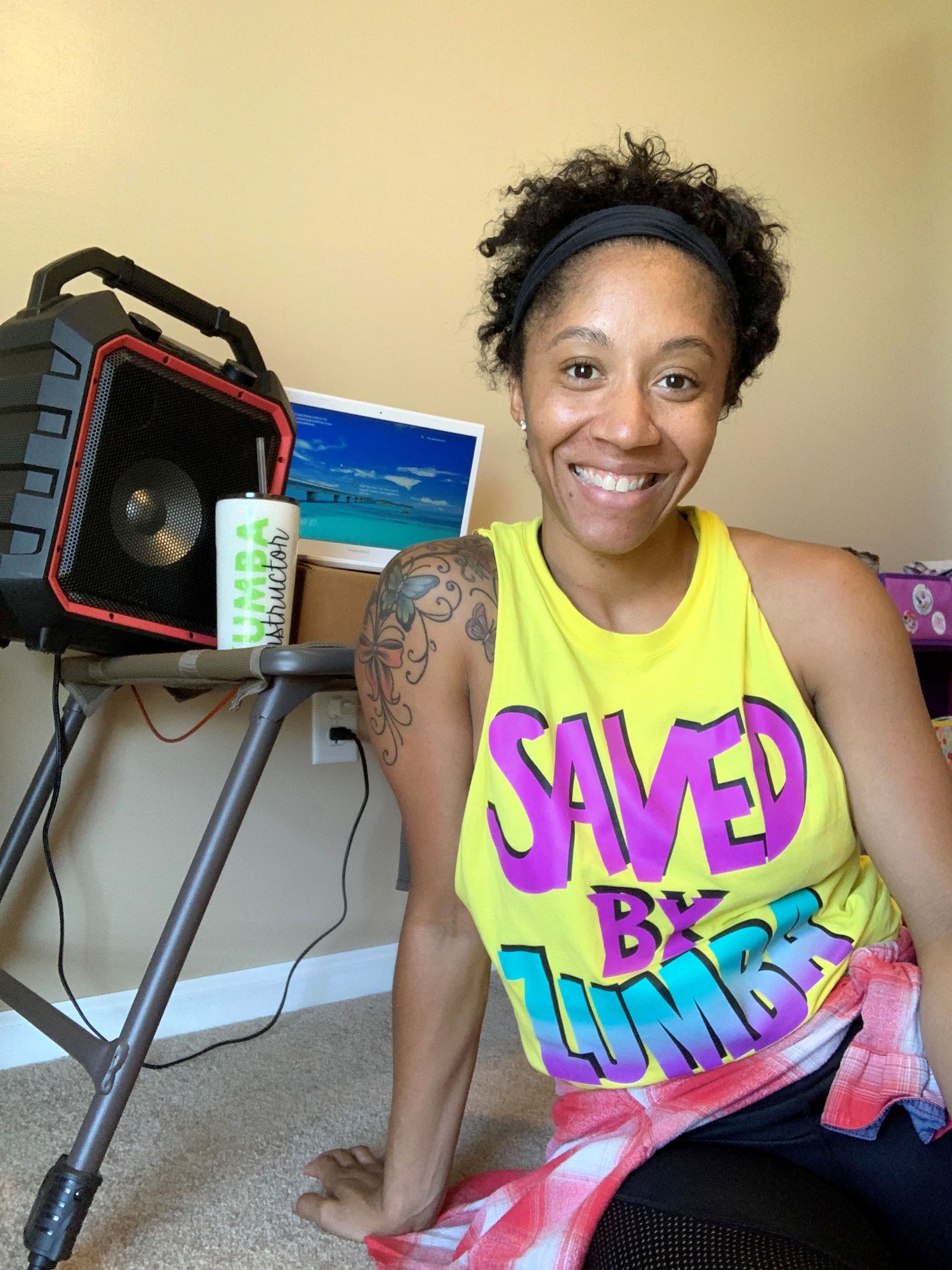 Photo shows a woman sitting in front of her tablet and music speaker smiling for the camera with the words, "Saved by Zumba" on her workout shirt.