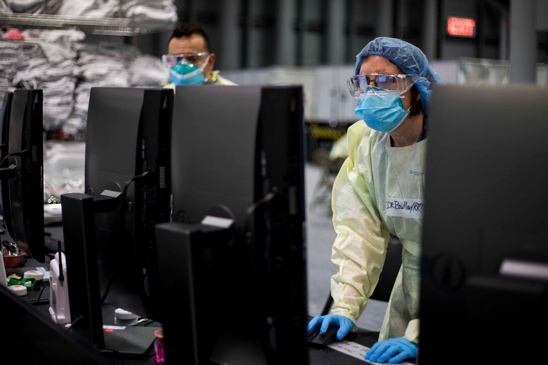 A sailor wearing personal protective equipment looks at computer monitors as another looks on.