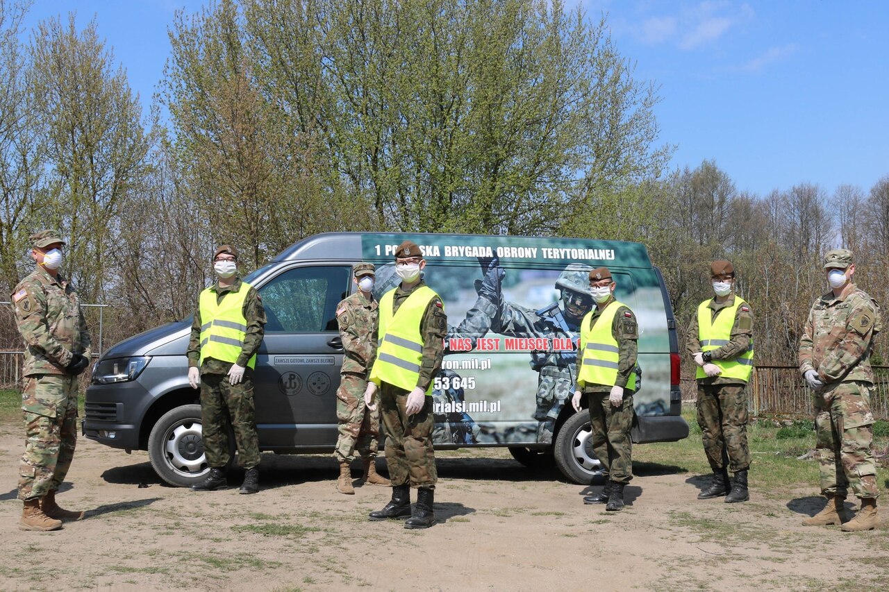 Uniformed military personnel also wearing personal protective equipment stand in front of a van.