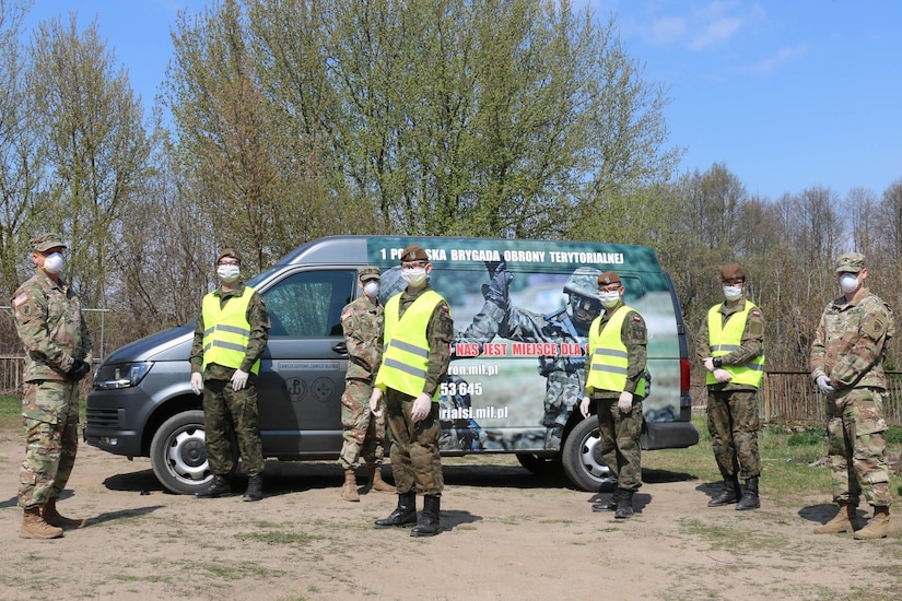 Uniformed military personnel also wearing personal protective equipment stand in front of a van.